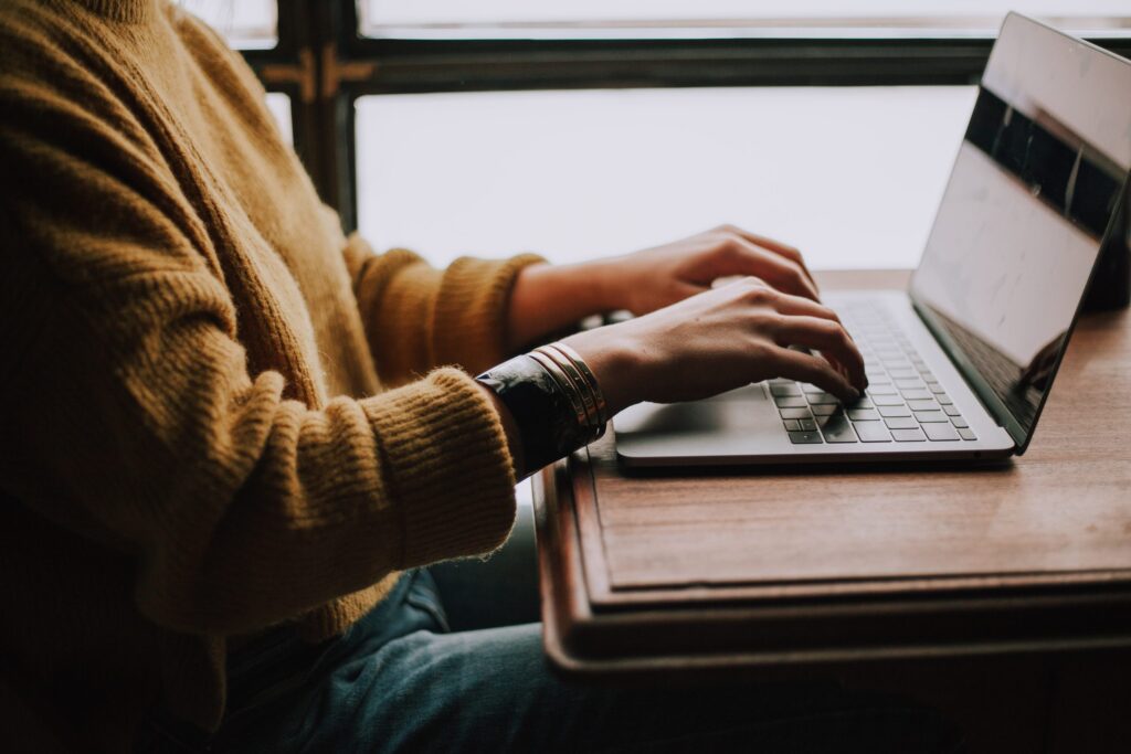 a woman wearing a yellow wool jumpers sits at a wooden table typing on a laptop